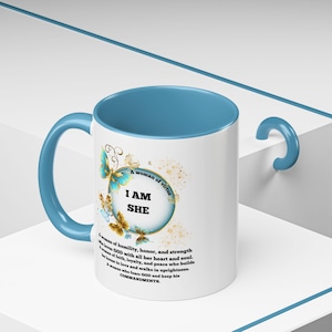 Könnte beinhalten: Eine weiße Keramiktasse mit hellblauem Rand und Henkel. Die Tasse zeigt eine Grafik eines Kreises mit Schmetterlingen und dem Text "A woman of virtue I AM SHE" sowie eine Beschreibung einer tugendhaften Frau.