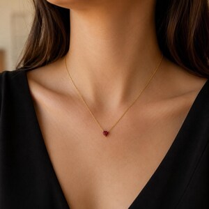 Peut inclure: Un collier à fine chaîne dorée agrémenté d'un petit pendentif en forme de cœur rouge profond. Le bijou repose sur la clavicule, mettant en valeur une chaîne en métal poli et une unique pierre précieuse centrale de couleur cramoisie.