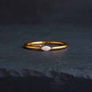 Peut inclure: Une fine bague en or dotée d'une pierre d'opale unique taillée en marquise, sertie horizontalement au centre. Le métal présente une finition polie et la pierre blanche irisée révèle de subtils reflets colorés sur le métal doré.