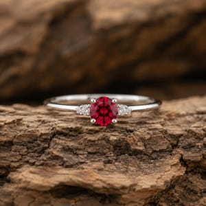 Peut inclure: Une bague en argent ornée d'une pierre précieuse ronde, facettée et rouge foncé au centre, flanquée de deux petites pierres d'accent rondes et transparentes de chaque côté, le tout sur un anneau en métal poli au fini lisse et brillant.
