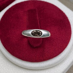 Peut inclure: Une bague en argent poli dotée d'un anneau lisse et effilé, sertie d'une pierre précieuse ovale de couleur brun foncé au centre. Le métal présente une finition métallique réfléchissante et la pierre offre un aspect facetté brun profond.