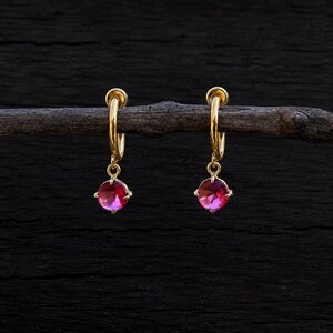 Peut inclure: Une paire de boucles d'oreilles créoles en or avec une petite pierre précieuse rose ronde à facettes. Chaque boucle se compose d'un anneau en or fin avec une pierre rose circulaire suspendue, présentant une surface brillante et polie.