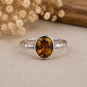 Peut inclure: Une bague en argent avec une pierre précieuse brune de taille ovale sertie dans un chaton lisse. L'anneau de la bague présente une texture martelée distincte, offrant une finition mate qui contraste avec la surface facettée et réfléchissante.