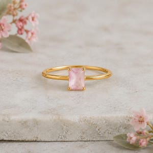 Peut inclure: Une bague en or ornée d'une pierre précieuse rectangulaire rose clair, fixée par un sertissage à quatre griffes. L'anneau est fin, lisse et poli, présentant un design épuré qui met en valeur la pierre rose pâle et translucide au centre.