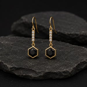 Peut inclure: Une paire de boucles d'oreilles pendantes dorées avec une barre verticale ornée de quatre petites pierres claires, reliée à un pendentif hexagonal en pierre noire. Les boucles d'oreilles ont une attache à crochet et une finition métallique dorée.