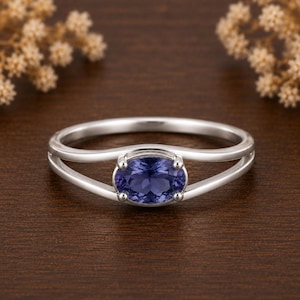 Peut inclure: Une bague argentée avec une pierre précieuse ovale d'un violet profond fixée par quatre griffes. L'anneau est divisé en deux brins métalliques fins et lisses qui convergent sur les côtés, créant un espace ouvert autour de la pierre facettée.