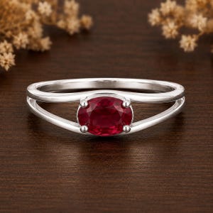 Könnte beinhalten: Ein silberfarbener Ring mit einem zentralen, oval geschliffenen, tiefroten Edelstein, der von vier Krappen gehalten wird. Die Ringschiene ist in zwei parallele Abschnitte unterteilt, die an den Seiten zusammenlaufen und einen offenen Raum bilden.