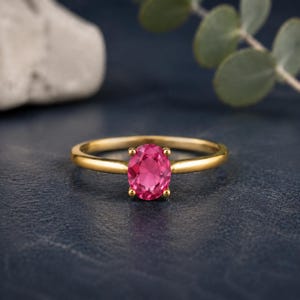 Peut inclure: Une bague en or ornée d'une pierre précieuse rose vif de taille ovale, fixée par quatre griffes. L'anneau est lisse et poli, présentant un design classique qui met en valeur la surface facettée et la couleur magenta profonde de la pierre.