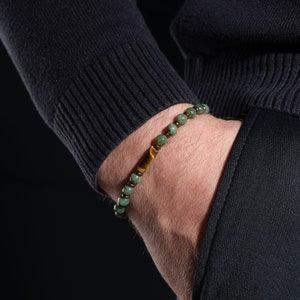 Puede incluir: Una pulsera de cuentas con cuentas verdes y marrones y un acento de ojo de tigre. La pulsera se lleva en la muñeca, con el brazo y la mano de la persona visibles. El fondo es oscuro.