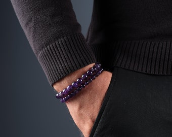 May include: Two amethyst beaded bracelets on a wrist. The bracelets are made of round, polished purple gemstones. The person is wearing a black jumper and black trousers, with their hand in their pocket.