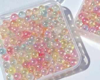 May include: Two clear plastic containers filled with iridescent pastel-colored beads. The beads are small and round, and they have a pearlescent finish. The containers are square and have lids.
