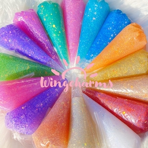May include: Assortment of 15 colorful glitter glue tubes in cone-shaped packaging. Each tube features a different color and glitter combination. The tubes are arranged in a circle with the brand name "Wingcharms" in the center.