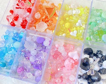 May include: Assortment of colorful plastic beads in clear containers. The beads are in various shapes, including hearts, stars, flowers, and geometric shapes. The beads are arranged in a rainbow pattern, with red, orange, yellow, green, blue, purple, pink, black, and white beads.