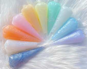 May include: A set of ten iridescent, glittery, cone-shaped containers filled with colorful slime. The containers are arranged in a circle on a white fluffy surface.