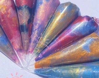 Könnte beinhalten: Ein Satz von acht schillernden Farbtuben mit einem galaxieartigen Wirbelmuster. Die Tuben sind fächerförmig angeordnet, wobei die Spitzen zum Zentrum zeigen. Die Farbe ist eine Mischung aus Rosa, Lila, Blau und Gold.