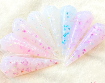 May include: A set of nine clear, cone-shaped tubes filled with a translucent, glittery slime. The slime is pink, white, and blue with small, iridescent hearts and stars. The tubes are arranged in a fan shape on a white, fuzzy surface.