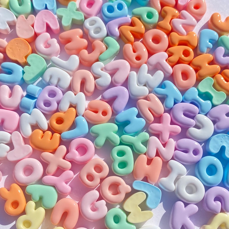 May include: A collection of colorful plastic letter beads in pastel shades of pink, blue, green, yellow, and white. The beads are shaped like letters of the alphabet and are perfect for crafting projects.