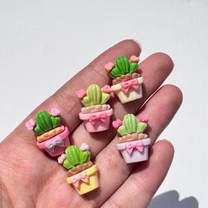 May include: Miniature cactus figurines in various pastel colors. Each cactus is potted in a different colored pot, with a pink bow and small pink flowers. The cacti are green and light green, with a brown base.