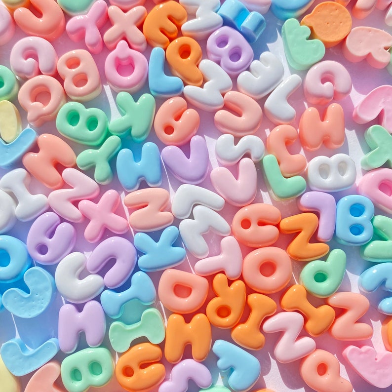 May include: A collection of colorful plastic letter beads in various pastel shades, including pink, blue, green, yellow, and orange. The beads are shaped like uppercase letters of the alphabet.