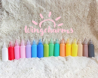 May include: A row of colorful squeeze bottles with white tips, arranged on a fluffy white surface. The bottles are filled with various shades of paint, including red, pink, blue, green, and yellow. The text "wingcharms" is written in pink.