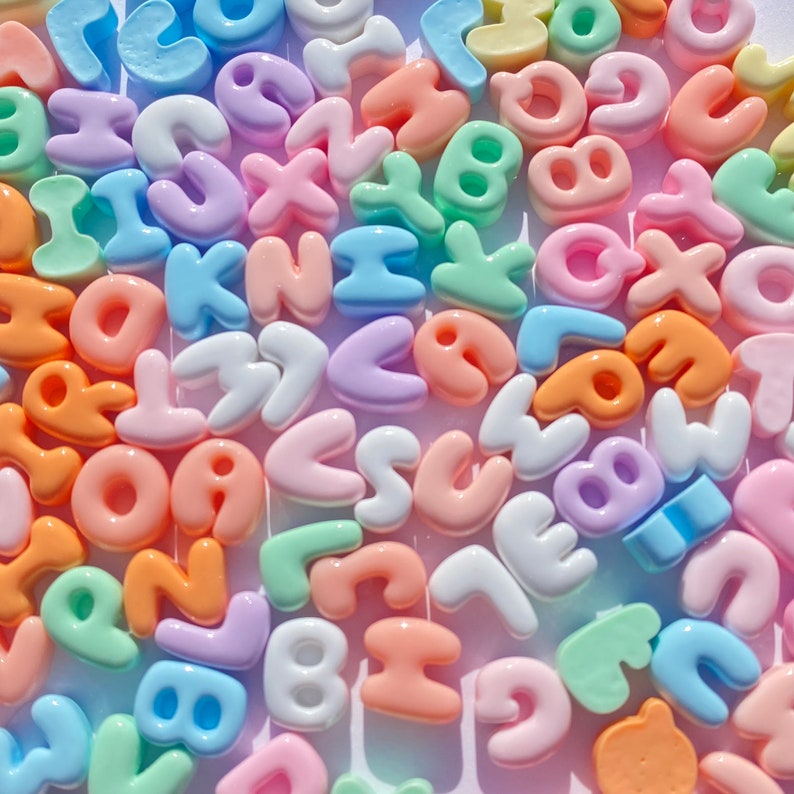 May include: A collection of colorful plastic letter beads in pastel shades of pink, blue, green, and yellow. The beads are shaped like letters of the alphabet and are perfect for crafting projects.