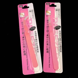 Puede incluir: Dos pinzas de depilar rosas empaquetadas en cart&oacute;n blanco y rosa. El empaque incluye texto japon&eacute;s que dice "anti&aacute;cido y no corrosivo Acero inoxidable" y "resistente al magnetismo y a la oxidaci&oacute;n".
