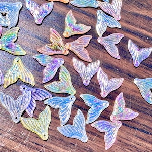 May include: A selection of iridescent mermaid tail charms in shades of blue, purple, pink, and yellow. These decorative charms have a shimmering, glittery effect and are scattered on a wooden surface.