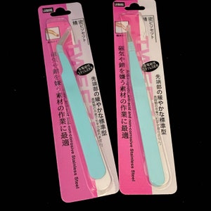 Puede incluir: Dos pares de pinzas de acero inoxidable azules con un embalaje rosa. El embalaje tiene texto japon&eacute;s que dice "Acero inoxidable antimagn&eacute;tico, anti&aacute;cido y no corrosivo" y "Para trabajos que requieren materiales sensibles al magnetismo, los &aacute;cidos y la corrosi&oacute;n."