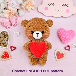 Può includere: Schema a uncinetto per un orsacchiotto marrone che tiene un cuore rosso. Lo schema è scritto in inglese ed è disponibile per il download in PDF. L'immagine include anche diverse decorazioni a forma di cuore.