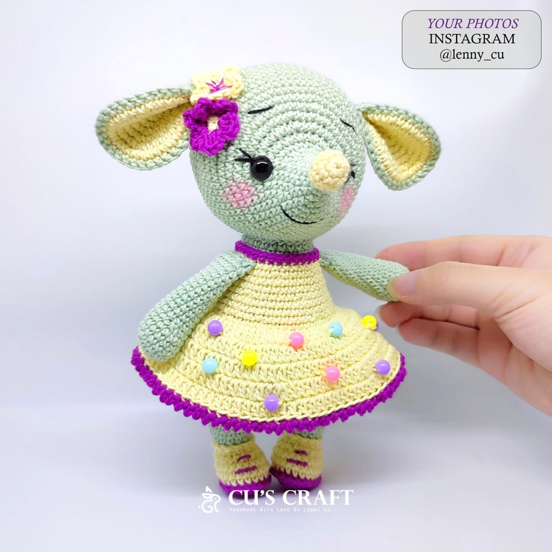 May include: A crocheted green and yellow doll with a purple flower on its head. The doll is wearing a yellow dress with a purple trim and colorful beads. The doll is standing on a white surface.  YOUR PHOTOS INSTAGRAM @lenny_cu