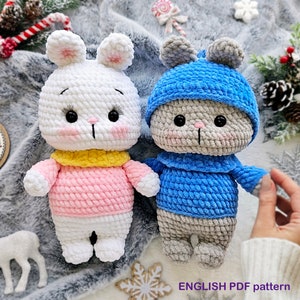 Peut inclure: Deux animaux en peluche crochetés, un lapin blanc portant un pull rose et un lapin gris portant un chapeau et un pull bleus. Les lapins sont assis sur une couverture moelleuse grise et blanche avec des flocons de neige et un arrière-plan sur le thème de Noël.