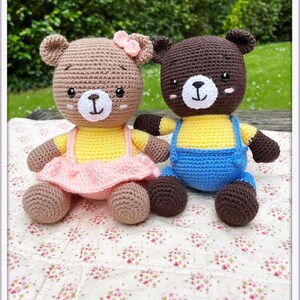 May include: Two crocheted teddy bears, one brown with a pink dress and a yellow shirt, the other brown with a blue overall and a yellow shirt, sitting on a white floral blanket. The text "Bear Polly and bear Nick" is above the bears.