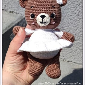 May include: A brown crocheted teddy bear wearing a white crocheted dress and veil. The bear has black eyes and a pink nose. The bear is being held by a hand.