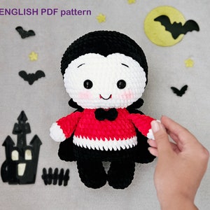 Puede incluir: Un juguete de peluche de crochet que representa a un vampiro con una camisa a rayas rojas y blancas, pantalones negros y una capa negra. El juguete tiene una cara blanca con ojos negros, una nariz roja y una gran sonrisa.