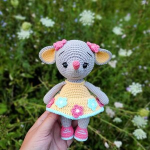 May include: A crocheted gray mouse wearing a yellow dress with pink and blue flowers. The mouse is holding a pink flower in its mouth and has pink bows on its ears.