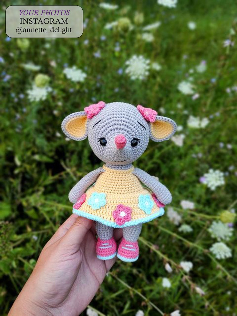 May include: A crocheted gray mouse wearing a yellow dress with pink and blue flowers. The mouse is holding a pink flower in its mouth and has pink bows on its ears.