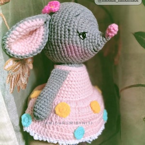 May include: A crocheted gray elephant wearing a pink dress with yellow and blue polka dots. The elephant has a pink flower on its head and a pink nose. The dress has a white lace trim.  YOUR PHOTOS INSTAGRAM @monda_handmade