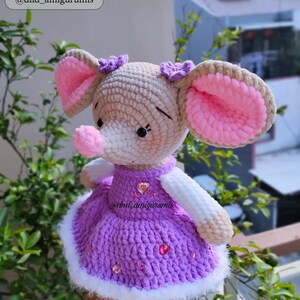 May include: A crocheted plush mouse wearing a purple dress with white trim and pink flowers on its head. The mouse has pink ears and a pink nose. The dress is decorated with pink rhinestones. The mouse is holding a pink heart with a rhinestone in the center. The text "@dnd_amigurumis" is visible on the dress.