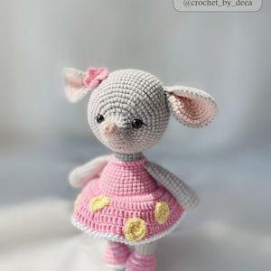 May include: A gray crocheted stuffed animal with pink ears and a pink dress with yellow polka dots. The animal has a pink flower on its head and black eyes.