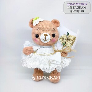 May include: A crocheted teddy bear wearing a white dress with lace trim and holding a bouquet of white flowers. The bear has a light brown body, black eyes, and a pink nose. The bear is wearing a small green flower on its head. The text "CU'S CRAFT" is visible in the bottom right corner of the image.