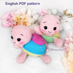 Pode incluir: Dois brinquedos de amigurumi de tartaruga de crochê rosa e branco com conchas coloridas. As tartarugas estão sentadas em uma superfície branca e fofa com flores rosa e brancas.