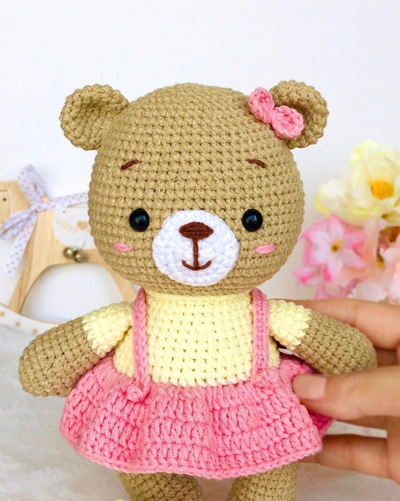 May include: A crocheted brown bear wearing a yellow and pink dress with a pink bow on its head.