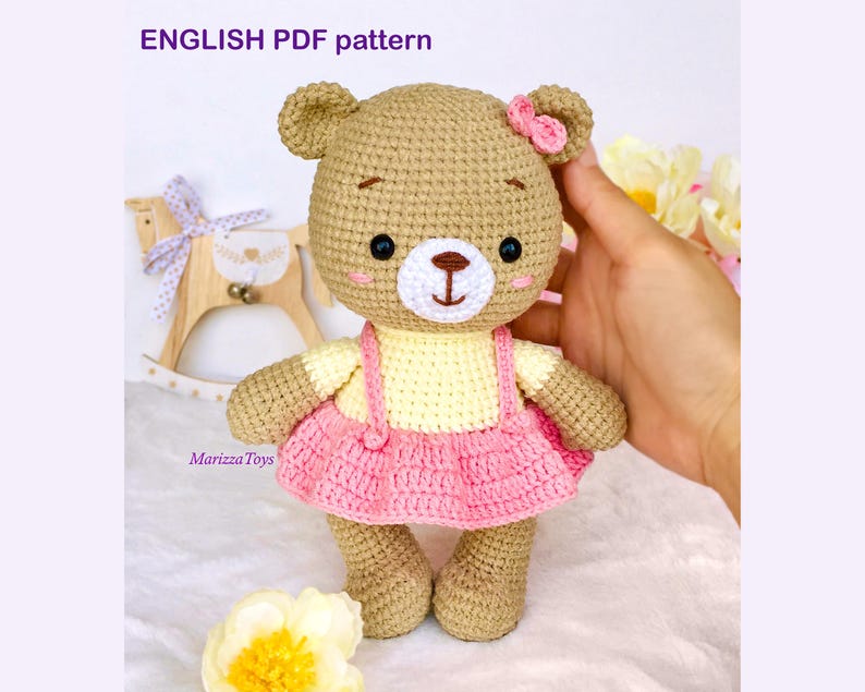 May include: Crochet pattern for a brown teddy bear wearing a pink dress and a yellow top. The pattern is available in English PDF format.