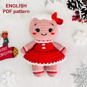 May include: A crocheted gingerbread girl doll wearing a red dress with white trim. The doll has a pink face with black eyes and a smiling mouth. The doll is sitting on a white background with snowflakes.
