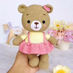 May include: A crocheted brown bear wearing a yellow and pink dress with a pink bow on its head.