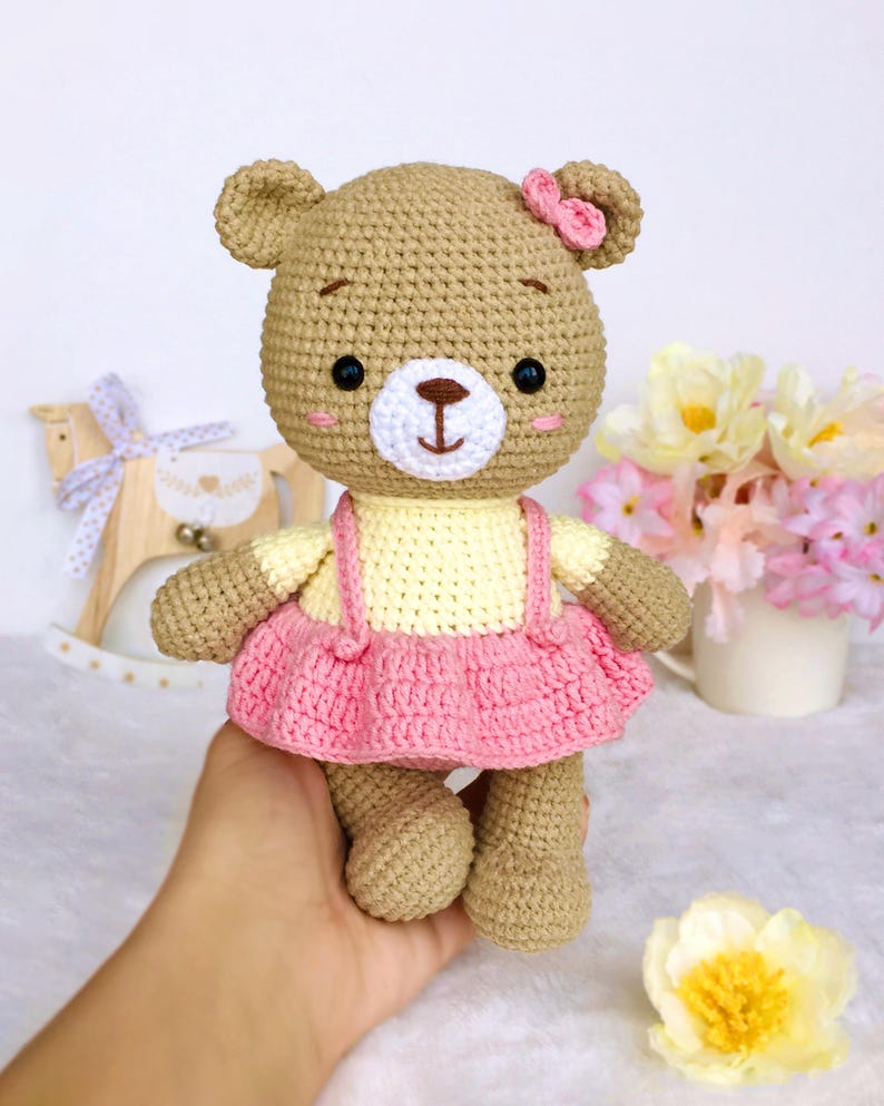 May include: A crocheted brown bear wearing a yellow and pink dress with a pink bow on its head.