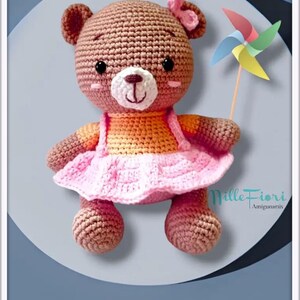 May include: A crocheted brown bear wearing a pink dress and an orange shirt, holding a colorful pinwheel. The bear has a pink bow on its head and black eyes. The bear is sitting on a light blue background. The text "Mille Fiori Amigurumi" is visible in the bottom right corner of the image.