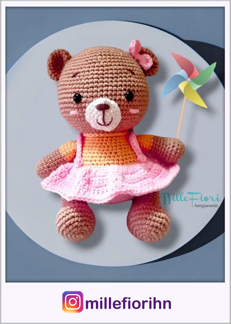 May include: A crocheted brown bear wearing a pink dress and an orange shirt, holding a colorful pinwheel. The bear has a pink bow on its head and black eyes. The bear is sitting on a light blue background. The text "Mille Fiori Amigurumi" is visible in the bottom right corner of the image.
