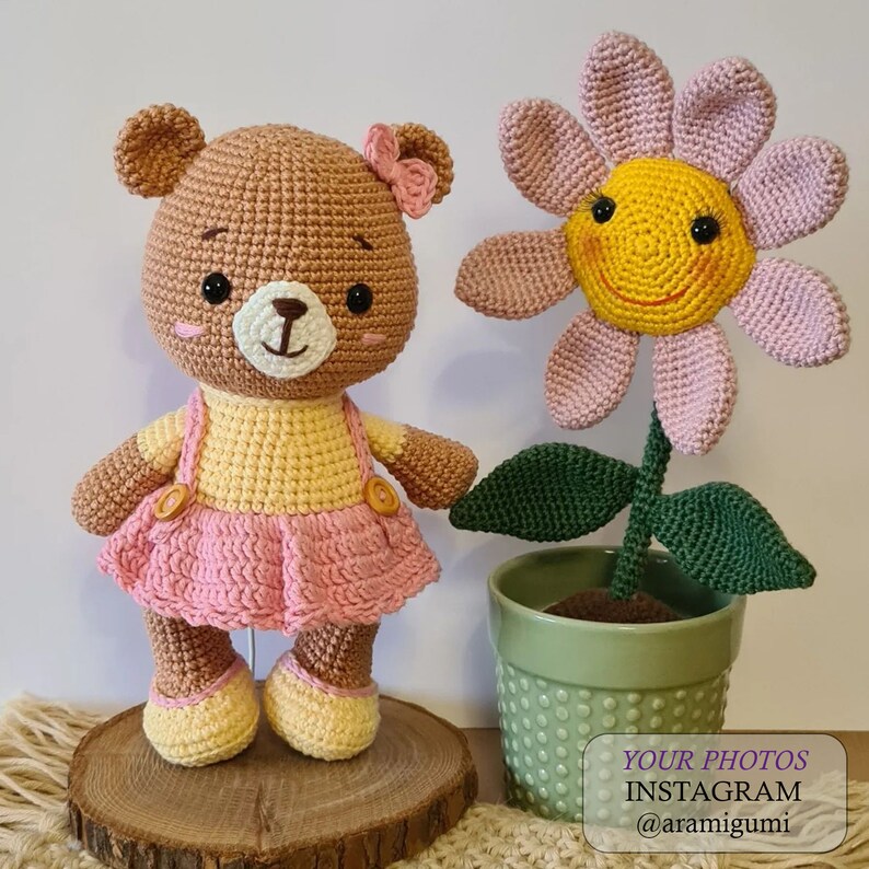 May include: A crocheted brown bear wearing a pink dress and a yellow top, standing next to a crocheted pink flower with a yellow smiley face. The flower is in a green pot with white dots. The text "YOUR PHOTOS INSTAGRAM @aramigurumi" is on a white background.