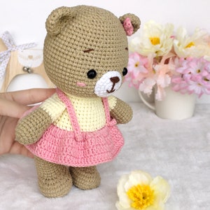 May include: A crocheted brown bear wearing a pink and yellow dress. The bear has black eyes and a white nose. The bear is standing on a white fluffy surface.